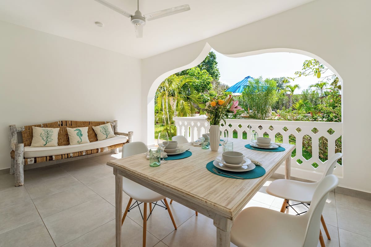 A dining area with a wooden table set for four, white chairs, a white sofa with decorative pillows, and a view of greenery through an arched window.