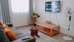 Ett vardagsrum med en soffa, ett soffbord, en tv som är monterad på väggen, en krukväxt och en golvfläkt.