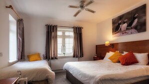 Une chambre d’hôtel avec deux lits simples, une tête de lit en bois, un ventilateur de plafond, une fenêtre avec des rideaux et un tableau représentant des macareux sur le mur.