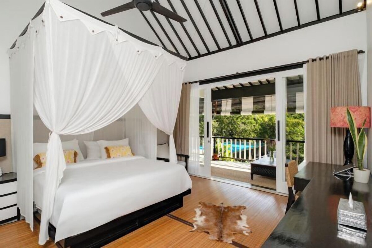A bedroom with a canopy bed, a wooden floor, a dining table, and a view of a pool and greenery.