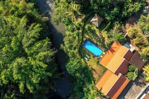 An aerial view of a property with a swimming pool surrounded by dense greenery, a small structure, and a building with a sloped roof. - Villa Lepak by the River (Bali)