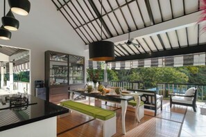 A modern kitchen with a central island, a dining area with a view, and a large window overlooking greenery. - Villa Lepak by the River (Bali)
