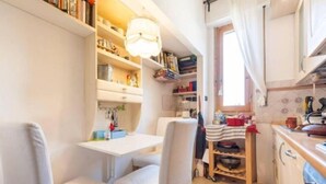 Une cuisine compacte avec des meubles blancs, une table à manger et des chaises. On y trouve des étagères remplies de livres et d’ustensiles de cuisine, ainsi qu’une fenêtre avec des rideaux.