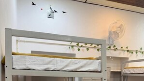 Une chambre avec un lit superposé, dotée de draps blancs et d’une couverture jaune. On y trouve un ventilateur de plafond et une décoration murale représentant des oiseaux et des cœurs.