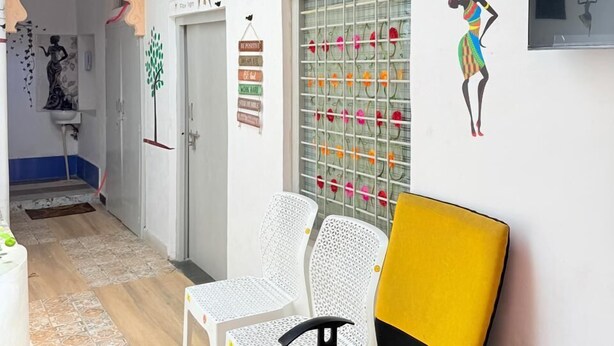 Une pièce avec un fauteuil jaune, des chaises blanches, un téléviseur, des œuvres d’art décoratives au mur et un sol à motifs.