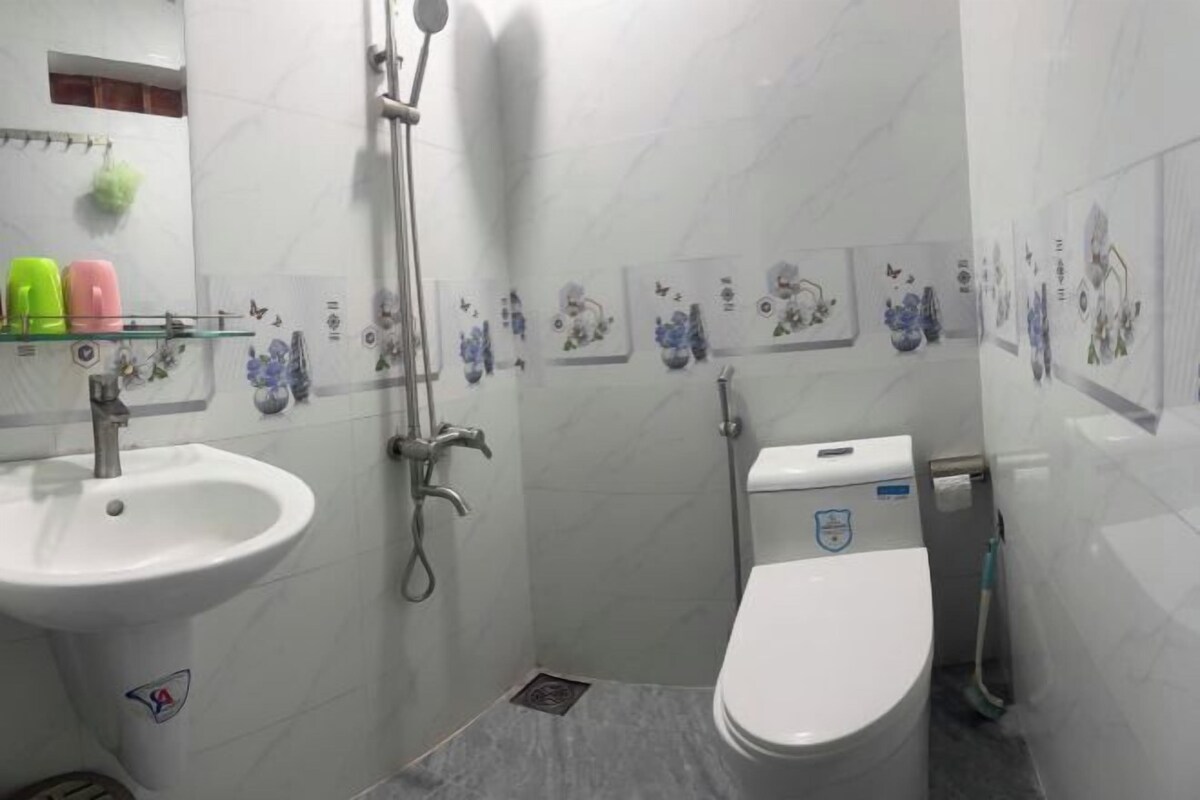 A small bathroom with a sink, toilet, and shower. There are decorative tiles on the walls and a small shelf with toiletries.