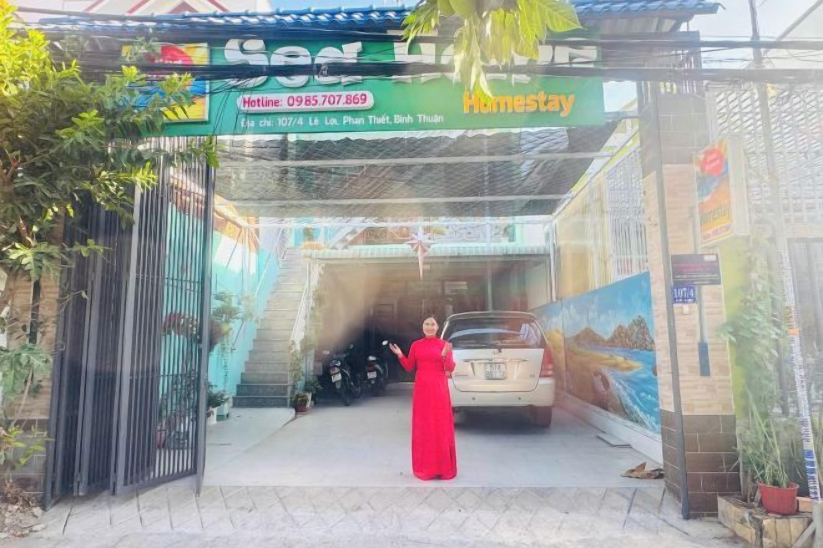 A woman in a red dress standing in front of a car at a property entrance with a sign that reads 'Homestay.'