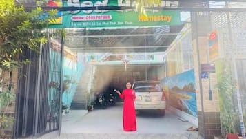 A woman in a red dress standing in front of a car at a property entrance with a sign that reads 'Homestay.'