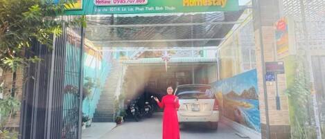 A woman in a red dress standing in front of a car at a property entrance with a sign that reads 'Homestay.'