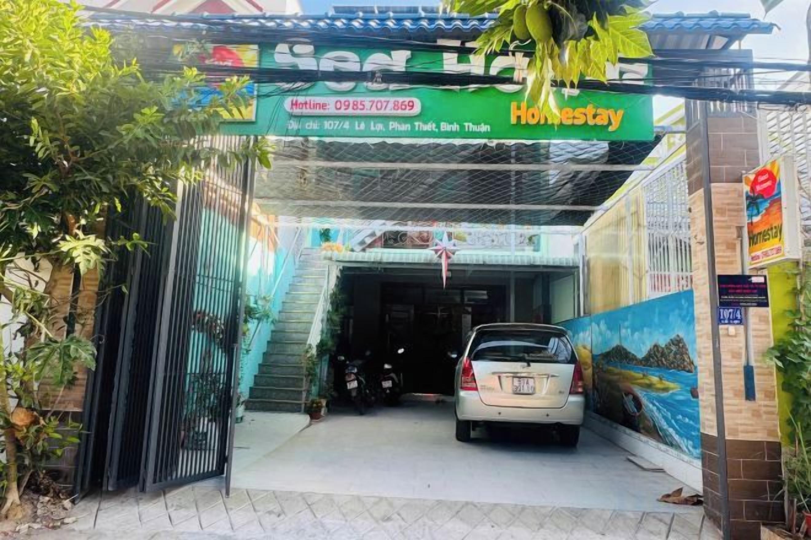 A hotel entrance with a car parked in front, a signboard with the hotel's name, and a contact number.