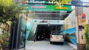 A hotel entrance with a car parked in front, a signboard with the hotel's name, and a contact number.