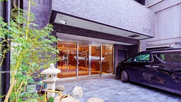 A modern building with a car parked in front, a stone pathway, and a small garden area with a stone lantern. - Shinsaibashi H2H 901 (Osaka)