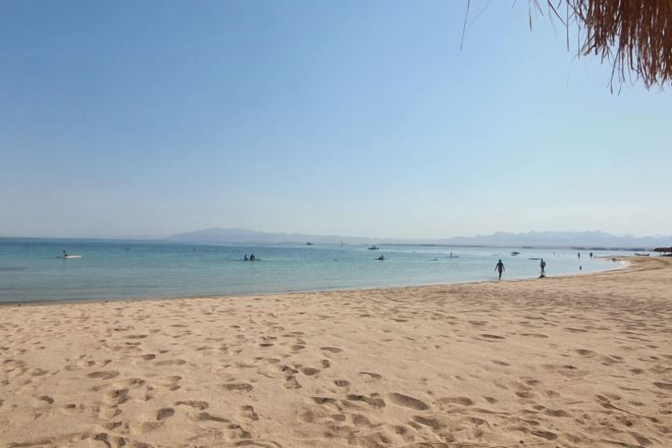 Uma praia com pegadas, águas calmas e barcos ao longe.