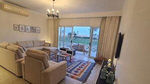 A living room with a sectional sofa, a coffee table, and a TV. There are large sliding glass doors leading to a balcony.