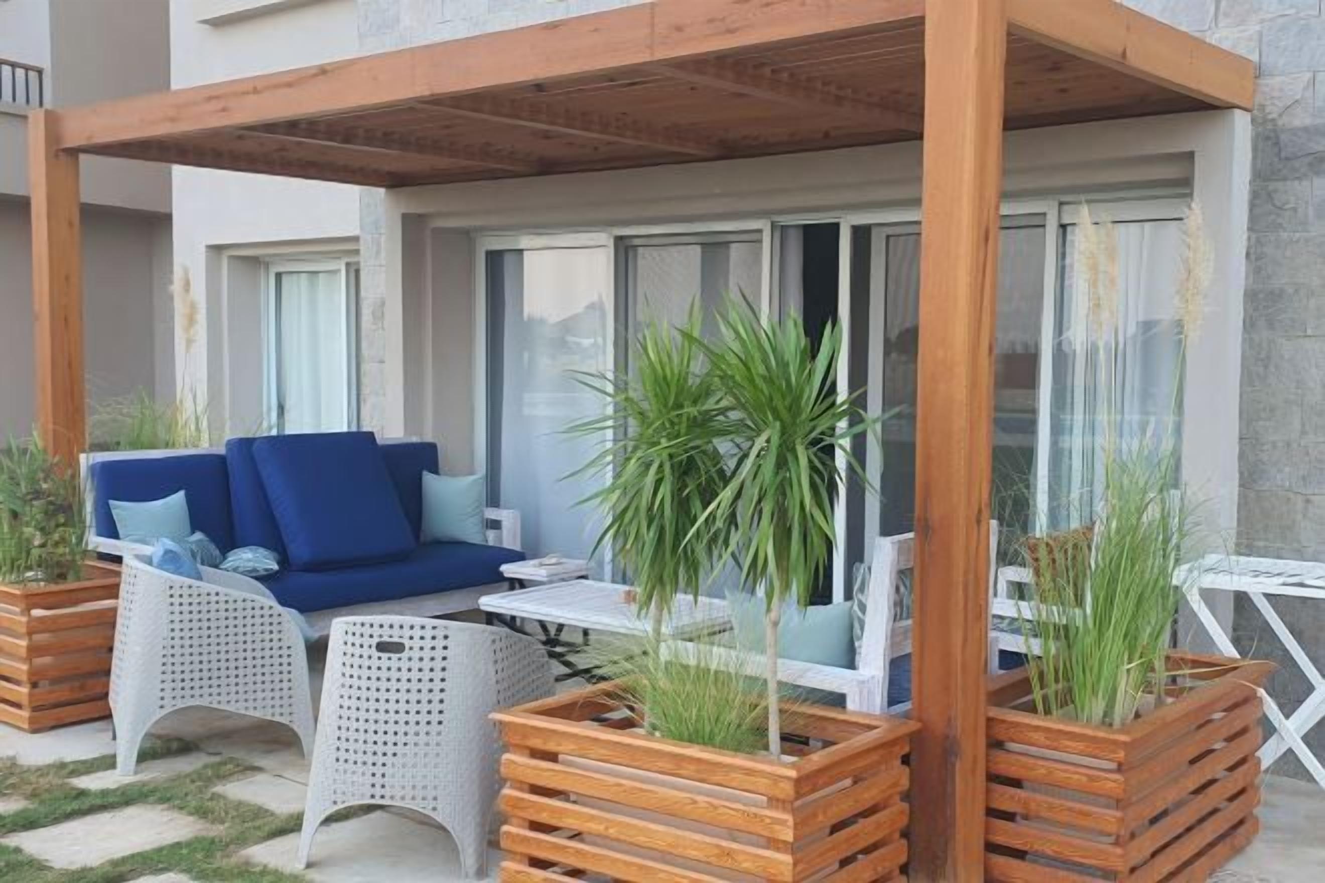 Un espacio de estar al aire libre con una pérgola de madera, muebles de mimbre blanco y plantas en macetas.