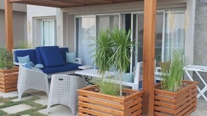 An outdoor seating area with a wooden pergola, a blue sofa, white wicker chairs, and wooden planters with greenery.