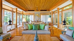 A spacious living room with a wooden ceiling, large windows, and a central sofa with green cushions. - The Sawan (Phuket)