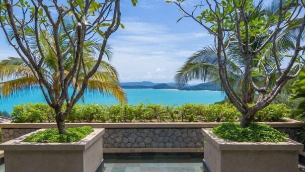A pool area with a view of a turquoise sea and mountains, surrounded by palm trees and planters. - The Sawan (Phuket)