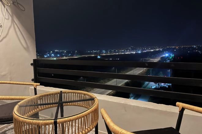 A balcony with a view of a cityscape at night, featuring a glass table and wicker chairs.