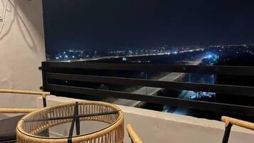 A balcony with a view of a cityscape at night, featuring a glass table and wicker chairs.