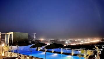 A rooftop pool with lounge chairs and umbrellas, overlooking a cityscape at night.