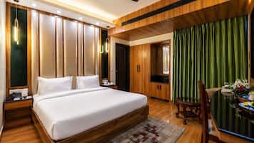 A hotel room with a large bed, wooden wardrobe, a desk with a chair, and a dining table with a green curtain backdrop.