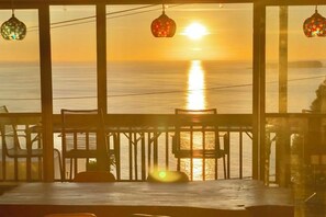 Une vue depuis un balcon, avec un coucher de soleil sur la mer, des chaises en bois et des lanternes suspendues.