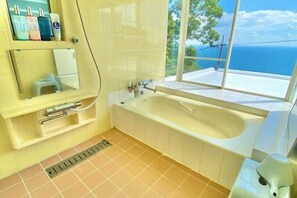 Une salle de bain avec une baignoire, une fenêtre donnant sur la mer et une étagère avec des articles de toilette.