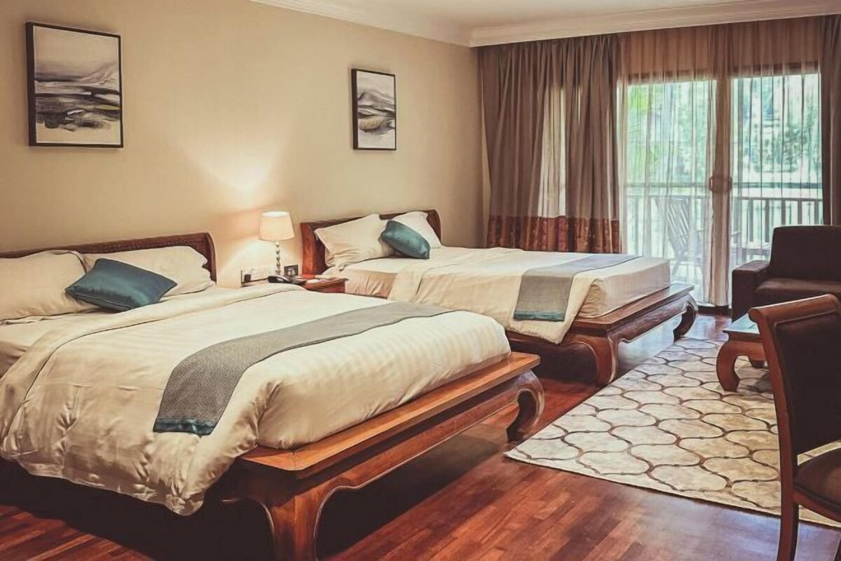 Ein Hotelzimmer mit zwei Betten, einem Holzkopfteil, einem Nachttisch, einer Lampe, einem Sessel und Blick ins Freie durch große Fenster.