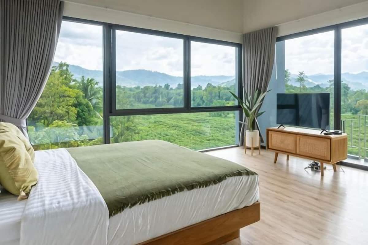 A bedroom with a large bed, a TV, a wooden cabinet, and a view of a lush landscape.