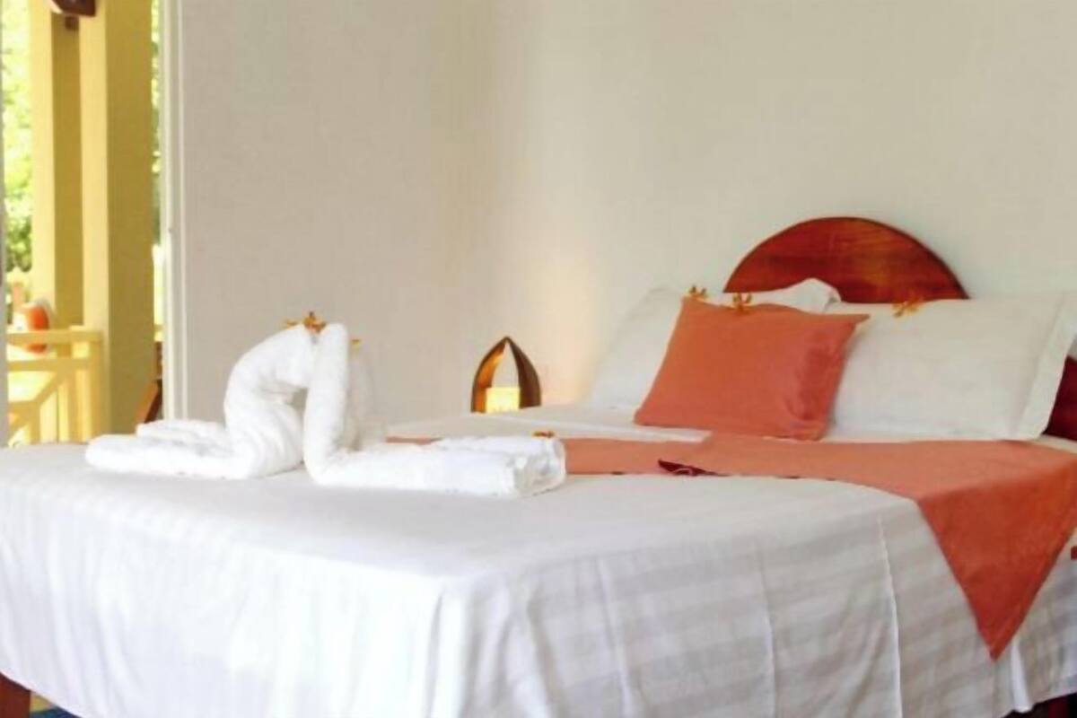 Ein ordentlich bezogenes Bett mit weiß-orangefarbener Tagesdecke, einem Holz-Kopfteil und einem Handtuch daneben.