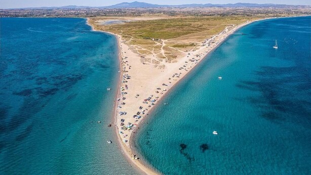 Vista aérea de una zona costera con playa, aguas azules cristalinas y montañas a lo lejos.
