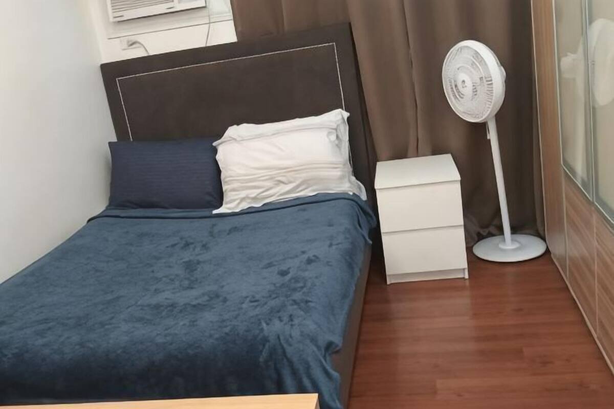 A single bed with a dark blue bedspread, a white bedside table, a white nightstand, and a standing fan.