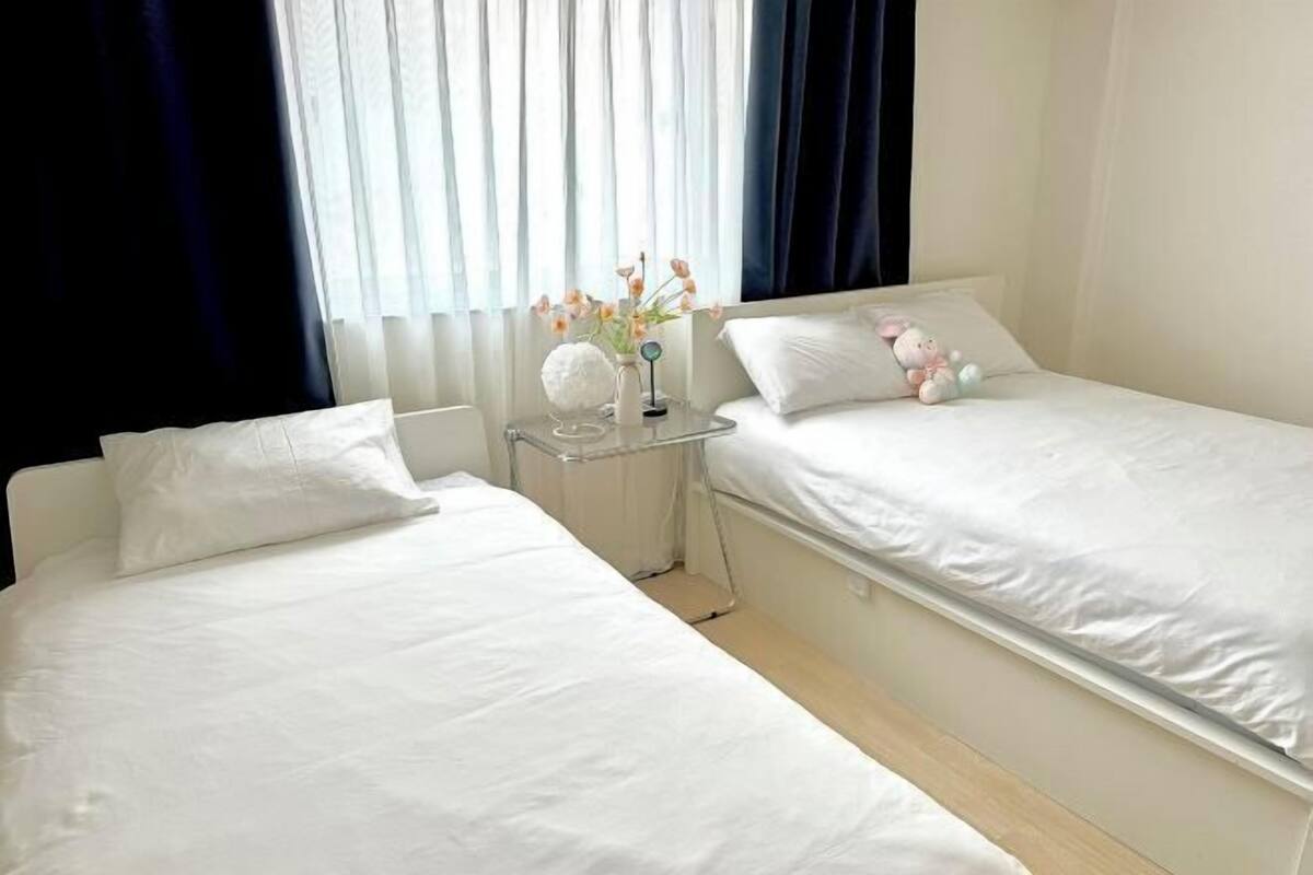 A hotel room with two single beds, a bedside table with a vase of flowers, and a window with curtains.