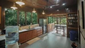 A modern kitchen with wooden cabinets, a water dispenser, and a refrigerator. There is a dining area with a table and chairs, and a bookshelf filled with books. - Deserenity Exclusive Jungle Resort Seremban (Seremban)