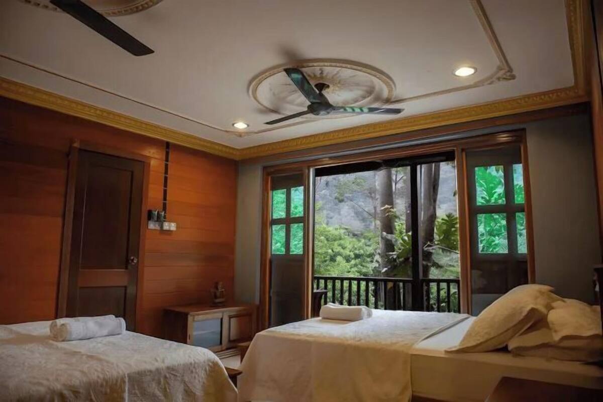 Habitación de hotel con dos camas, un ventilador de techo, una ventana con vistas a zonas verdes y una mesita de noche con lámpara.