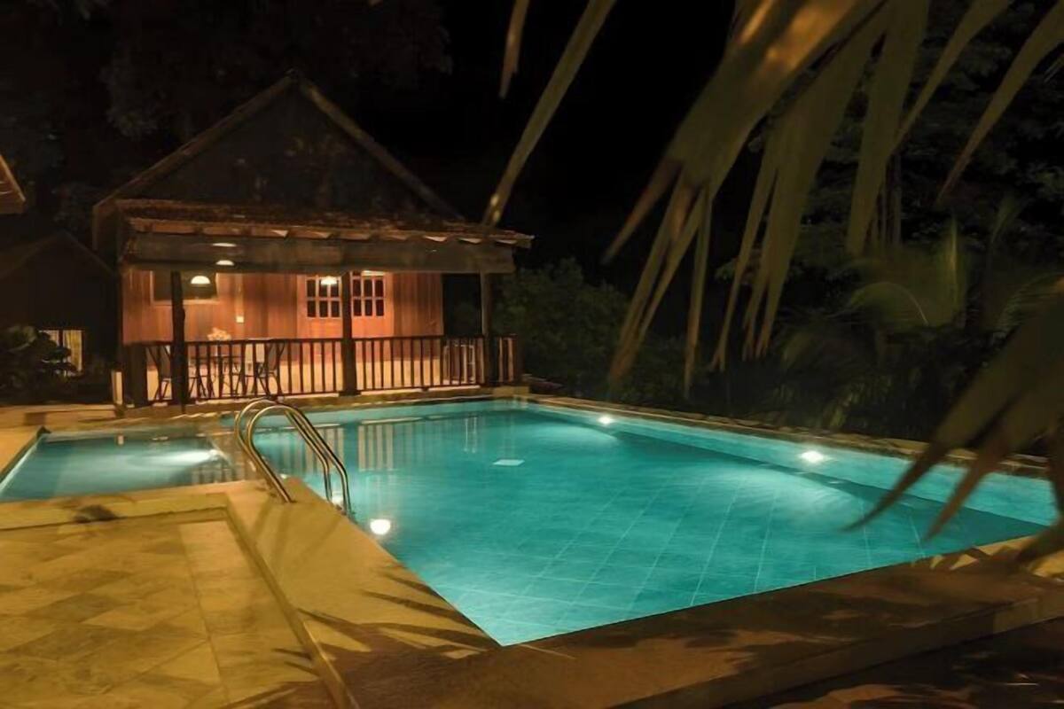 Una piscina con cubierta de madera y una casa pequeña al fondo.
