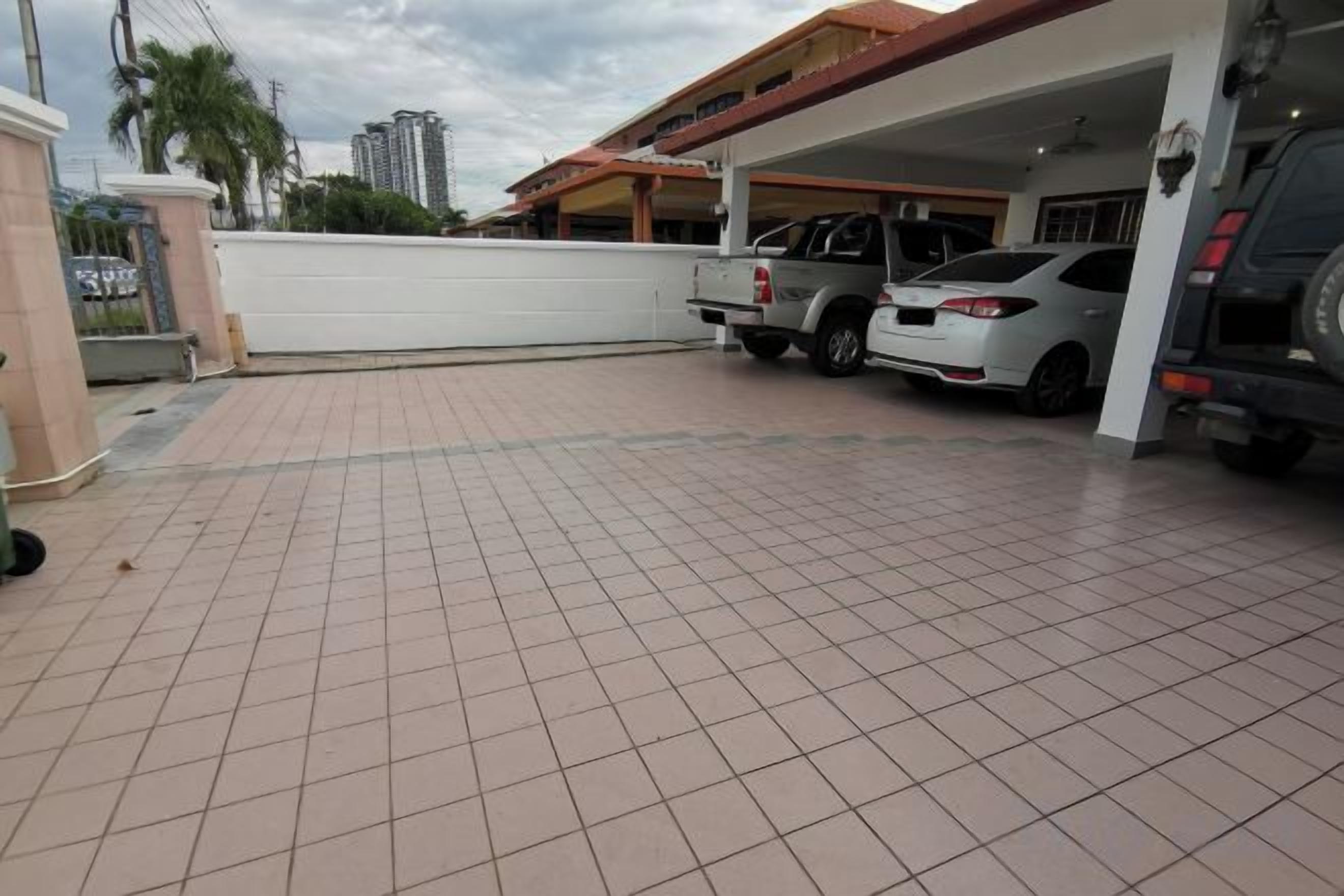 A covered parking area with a tiled floor, a white wall, and a building with a red roof in the background.