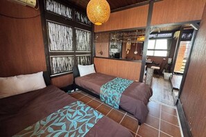 A room with two beds, a wooden wall, a hanging light fixture, and a view of the outside. - Nukanuka ぬかぬか (Miyakojima)