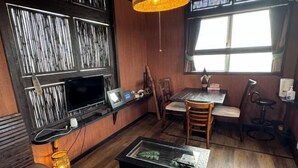 Une pièce avec un plancher en bois, une table à manger entourée de chaises, un téléviseur fixé au mur et une fenêtre avec des rideaux.