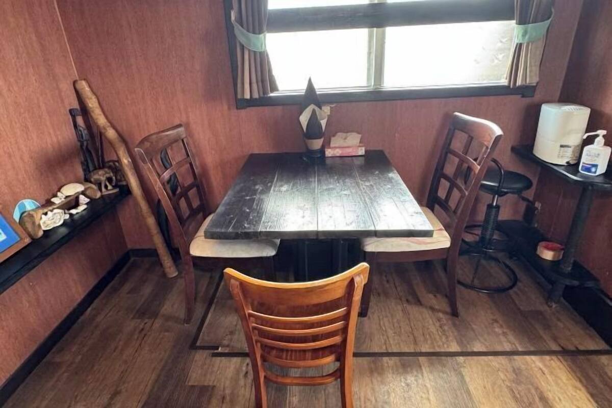 A wooden dining table with chairs, a window with curtains, and a small shelf with items.