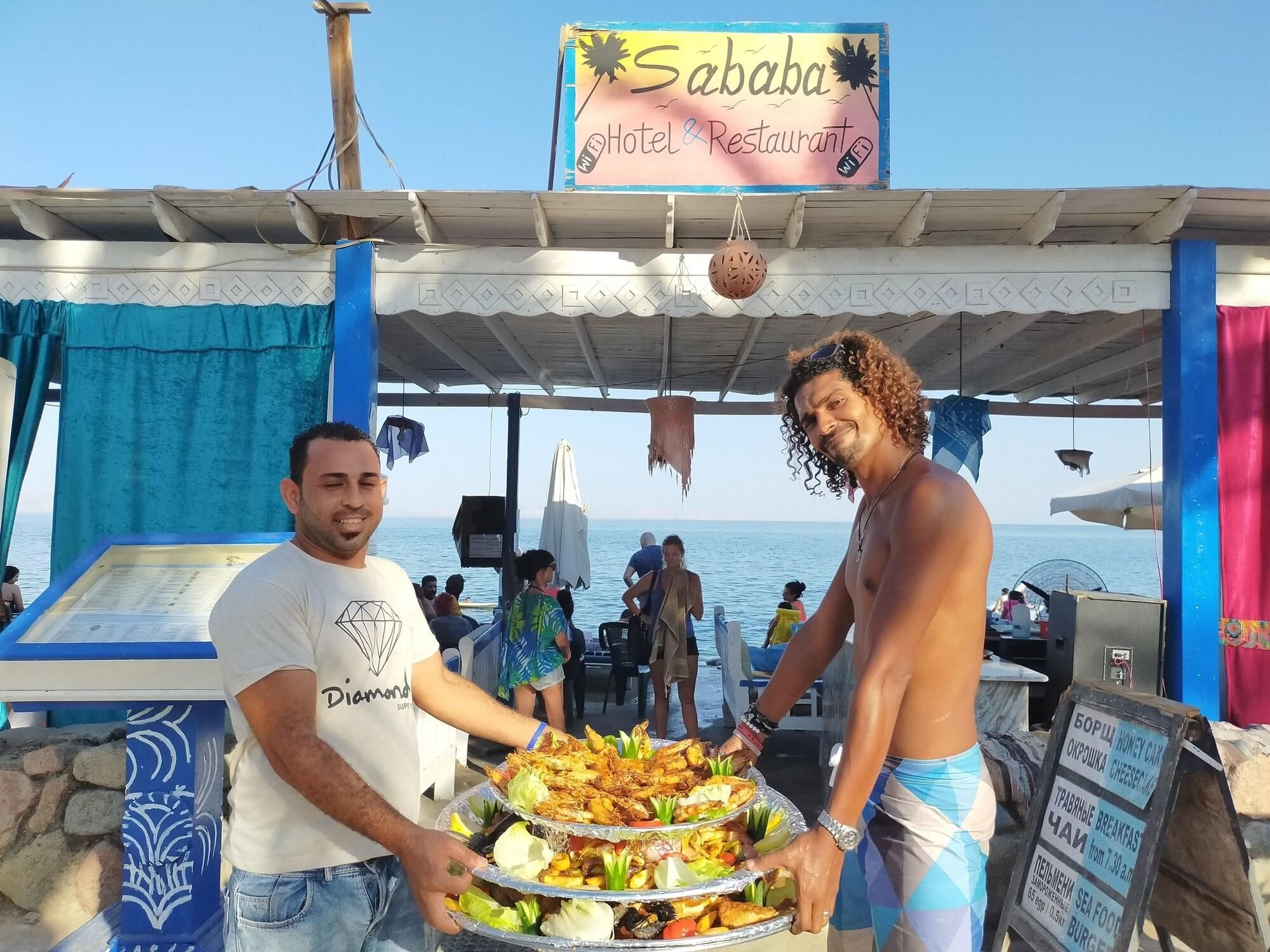 A seaside restaurant named 'Sababa' with a signboard, serving food on a table, and a menu board visible.