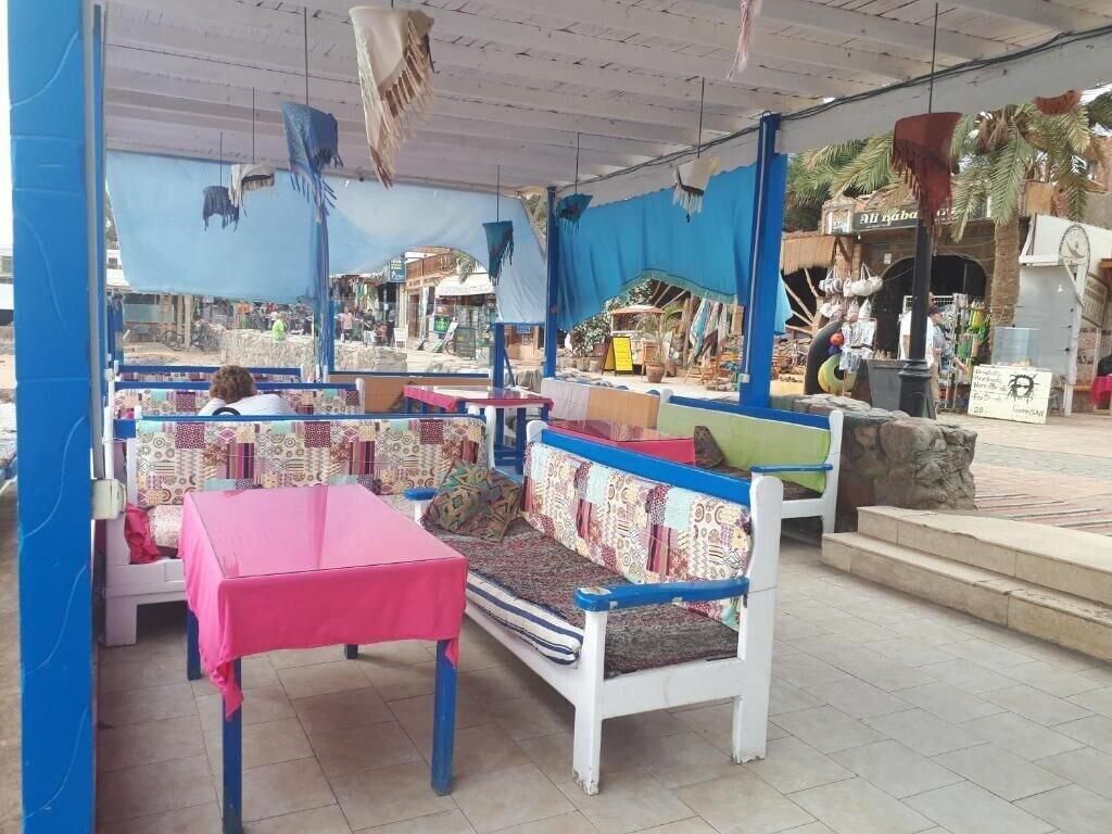 Un área de estar al aire libre con bancos y mesas, algunas cubiertas por lonas azules, y una fachada visible al fondo.