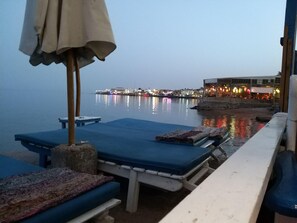 A waterfront view with lounge chairs and a table, a pier with buildings in the background, and a calm body of water reflecting lights. - Sababa Hotel Dahab (Dahab)