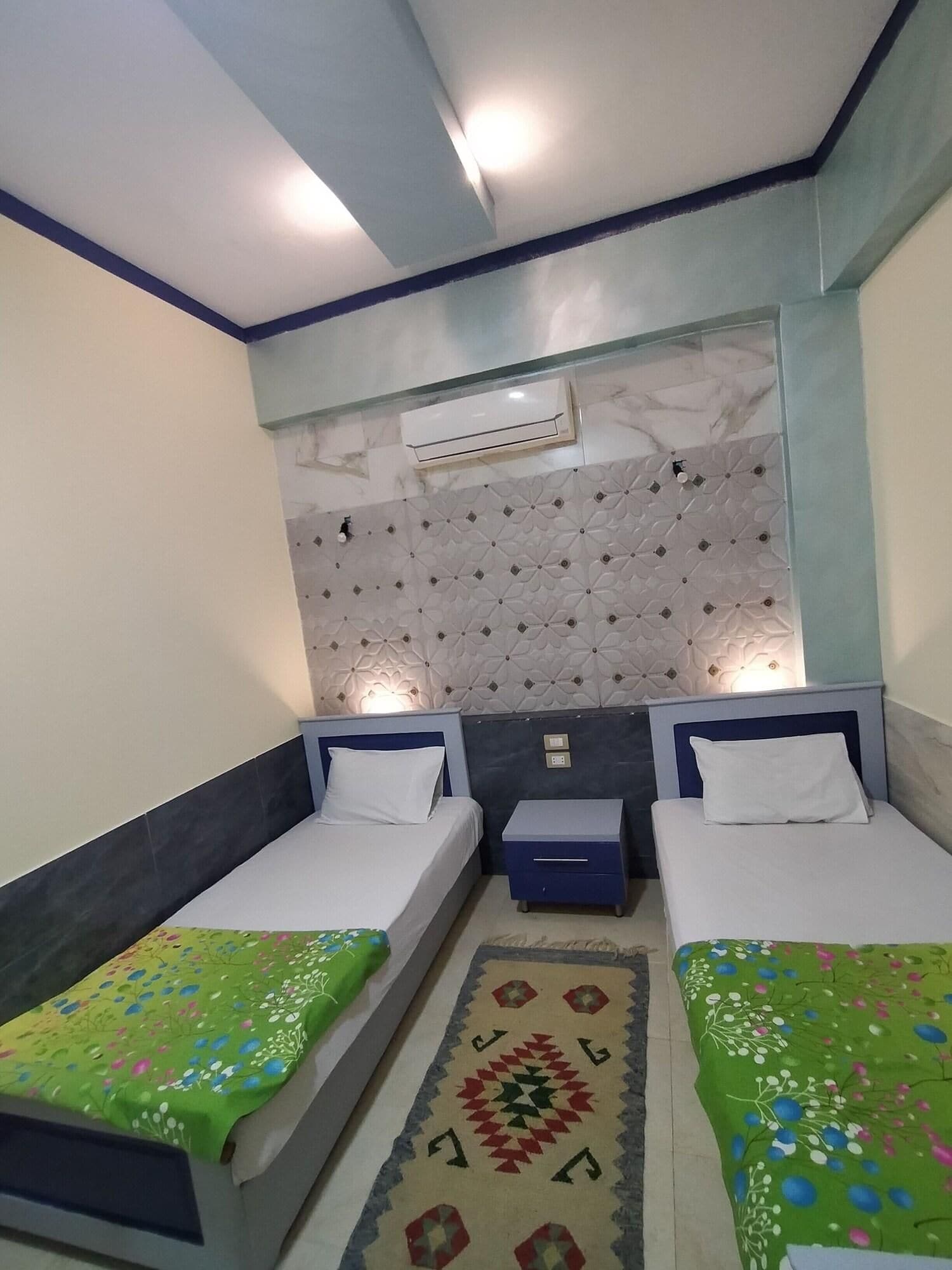 Quarto duplo com duas camas, mesa de cabeceira pequena, ar condicionado na parede e tapete decorativo.