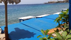 Blick auf das Meer mit einer Reihe blauer Liegestühle und einem weißen Sonnenschirm.