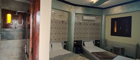 A hotel room with two beds, a shower, a small table, and a wall-mounted air conditioning unit.