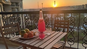 Een houten tafel met een fles Schweppes, een schaal fruit en twee glazen op een balkon met uitzicht op de ondergaande zon.