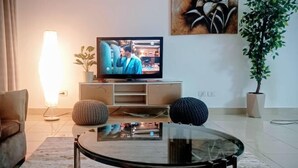 Een woonkamer met een televisie op een dressoir, een glazen salontafel, een bank, een lamp en twee potplanten.