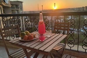 Una mesa de madera con una botella, un vaso y fruta en un balcón con vista al atardecer.